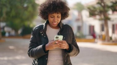 Parkta akıllı telefon kullanan neşeli yüzlü Afrikalı Amerikalı genç bir kadın.