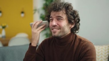 Genç İspanyol adam evdeki koltukta oturmuş akıllı telefondan sesli mesaj dinliyor.