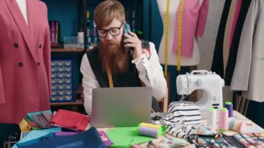 Young redhead man tailor talking on smartphone using laptop at clothing factory