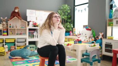 Young blonde woman preschool teacher stressed sitting on chair at kindergarten