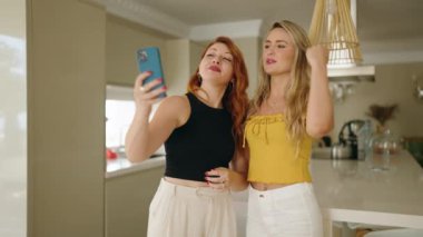 İki kadın akıllı telefondan selfie çekiyor. Evde yan yana duruyorlar.