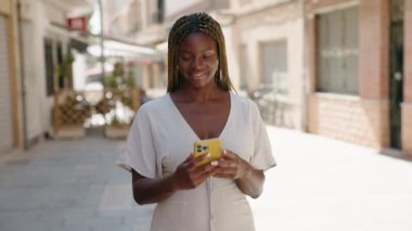 Afro-Amerikalı kadın gülümsüyor. Sokakta akıllı telefonun yanında özçekim yapıyor.