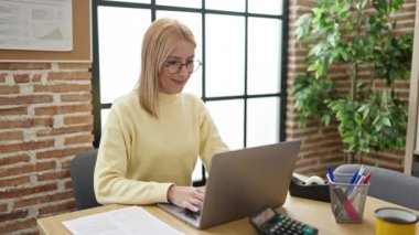 Genç sarışın iş kadını ofiste neşeli bir ifadeyle dizüstü bilgisayar kullanıyor.