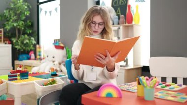 Young blonde woman preschool teacher reading book with relaxed expression at kindergarten