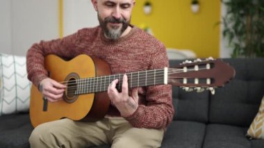 Genç kel adam evdeki kanepede oturup klasik gitar çalıyor.