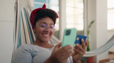 Ev terasındaki hamakta dizüstü bilgisayar ve kredi kartı kullanan Afrikalı Amerikalı kadın.
