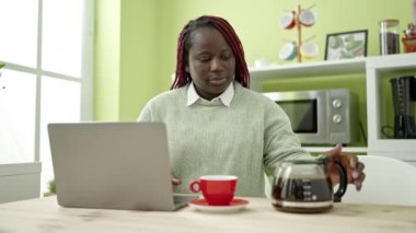Saçlarını örmüş, laptopunu kullanan, yemek odasında kahve içen Afrikalı bir kadın.