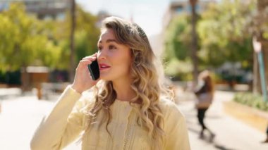 Genç sarışın kadın gülümsüyor. Parkta yürürken akıllı telefondan konuşuyor.