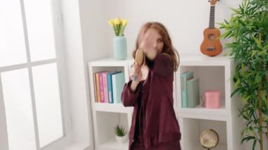 Young beautiful hispanic woman singing song using brush as a microphone at bedroom