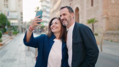 Erkek ve kadın çift birbirlerine sarılıyorlar. Sokağın karşısındaki akıllı telefonun yanında selfie çekiyorlar.