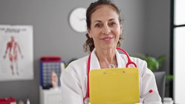 中年惊慌失措的女医生面带微笑 自信地在诊所拿着剪贴板 — 图库视频影像