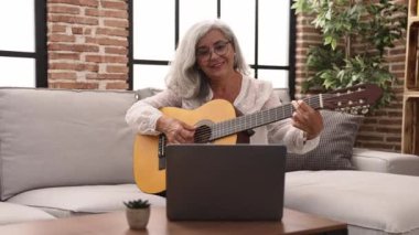 Middle age grey-haired woman smiling confident having online guitar class at home