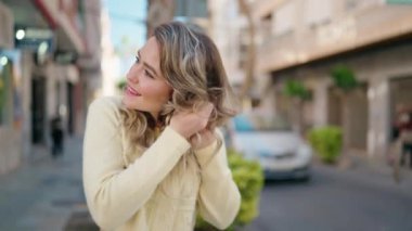 Young blonde woman smiling confident combing hair with hand at street