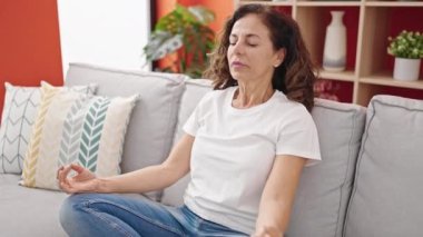 Orta yaşlı İspanyol kadın yoga egzersizi yapıyor. Evdeki kanepede oturuyor.