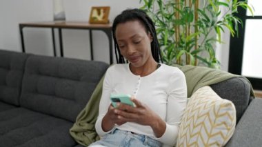 Evdeki koltukta oturan Afro-Amerikan kadın akıllı telefon kullanıyor