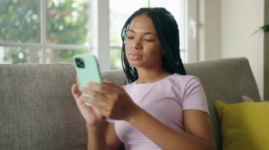 Evdeki koltukta oturan Afro-Amerikan kadın akıllı telefon kullanıyor