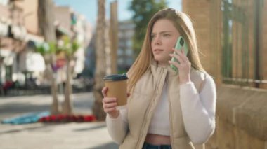 Genç sarışın kadın akıllı telefonda konuşuyor. Sokakta kahve içiyor.