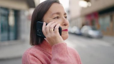 Orta yaşlı bir kadın gülümsüyor. Sokakta akıllı telefondan konuşuyor.