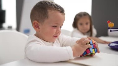 Adorable girl and boy playing with color puzzle cube sitting on table at kindergarten