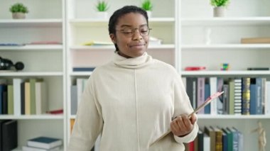 Afrikalı Amerikalı kız öğrenci kütüphane üniversitesinin panosunda kendinden emin bir şekilde kitap okurken gülümsüyor