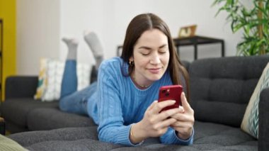 Beyaz kadın akıllı telefon kullanıyor. Evdeki kanepede yatıyor.