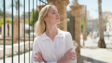 Genç sarışın kadın gülümsüyor. Kollarını kavuşturup caddede duruyor.