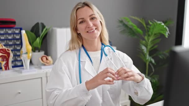 年轻的金发女医生微笑着在诊所用手做心脏手势 — 图库视频影像