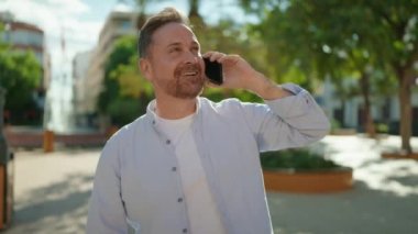 Genç beyaz adam gülümsüyor. Parktaki akıllı telefondan konuşuyor.