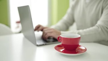 Genç İspanyol adam evde elinde bir fincan kahveyle dizüstü bilgisayar kullanıyor