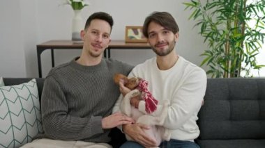 İki erkek, kanepede oturmuş, kendinden emin bir şekilde gülümsüyor ve evde köpeğe sarılıyorlar.