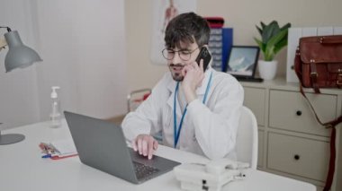 Genç İspanyol doktor bilgisayarını kullanarak klinikte telefonla konuşuyor.