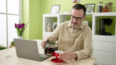 Orta yaşlı bir adam evde kahve içerken dizüstü bilgisayar kullanıyor.