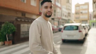 Genç Arap adam gülümsüyor. Kollarını kavuşturup caddede duruyor.