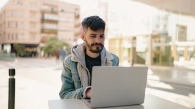 Young hispanic man using laptop sitting on table at coffee shop terrace