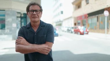 Orta yaşlı bir adam gülümsüyor. Kollarını kavuşturup caddede duruyor.