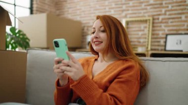 Genç kızıl saçlı kadın akıllı telefon kullanıyor. Yeni evindeki kanepede oturuyor.