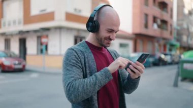 Genç kel adam sokakta akıllı telefon ve kulaklık kullanarak kendinden emin gülümsüyor.