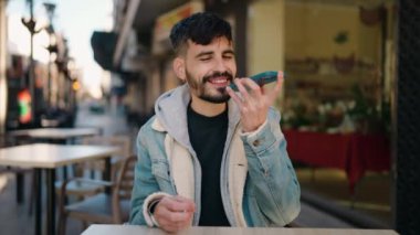 Young hispanic man talking on the smartphone sitting on table at coffee shop terrace
