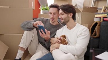 İki erkek akıllı telefon kullanıyor. Yeni evlerinde köpekleriyle yerde oturuyorlar.