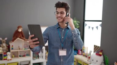 Young hispanic man preschool teacher using touchpad talking on smartphone at kindergarten