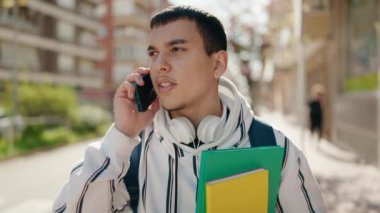 Young man student talking on the smartphone holding books at street