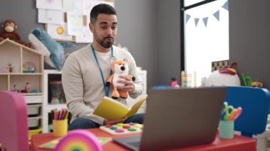 Young hispanic man preschool teacher reading story book having video call at kindergarten