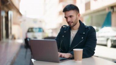 Young hispanic man using laptop sitting on table at coffee shop terrace