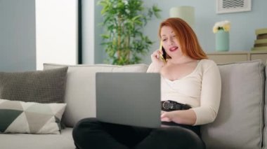 Genç kızıl saçlı kadın dizüstü bilgisayar kullanıyor ve evdeki akıllı telefondan konuşuyor.