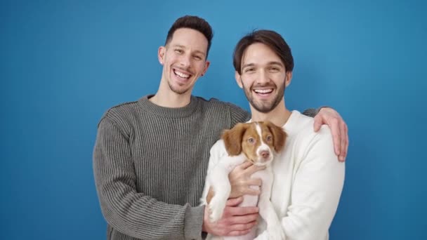 Двоє Чоловіків Посміхаються Впевнено Стоячи Обіймаючи Собаку Над Ізольованим Синім — стокове відео