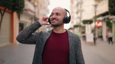 Genç kel adam gülerek sokakta müzik dinliyor.