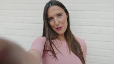 Genç, güzel, İspanyol bir kadın gülümsüyor. İzole edilmiş beyaz tuğla arka plan üzerinden görüntülü arama yaptırıyor.