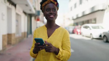 Genç Afro-Amerikalı kadın sokakta akıllı telefon kullanarak gülümsüyor.