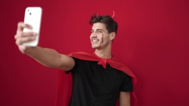 Şeytan kostümü giyen İspanyol genç adam izole edilmiş kırmızı arka planda akıllı telefondan selfie çekiyor.
