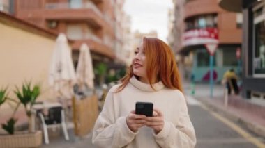 Caddede akıllı telefon kullanan, kendine güvenen genç kızıl saçlı kadın.